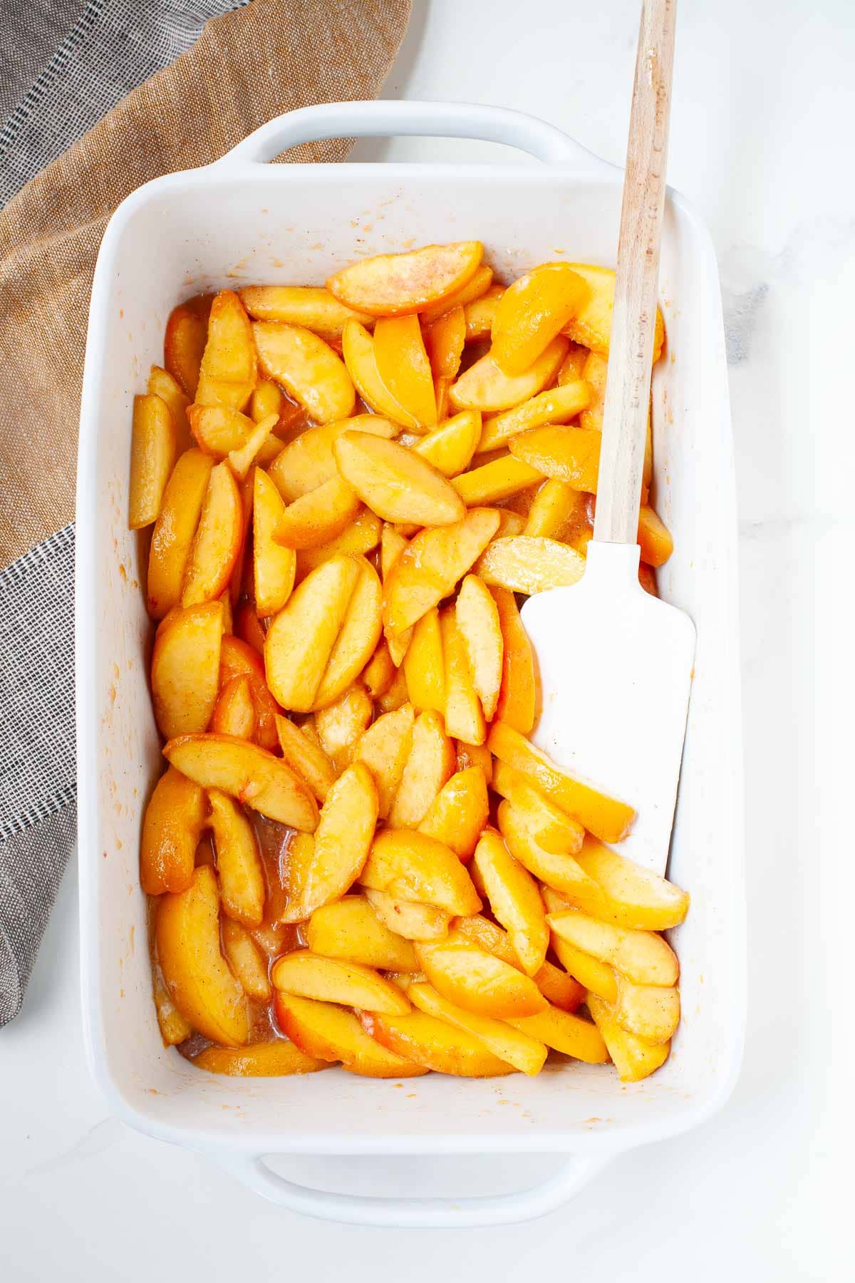 white baking dish filled with coated peaches and spatula on top