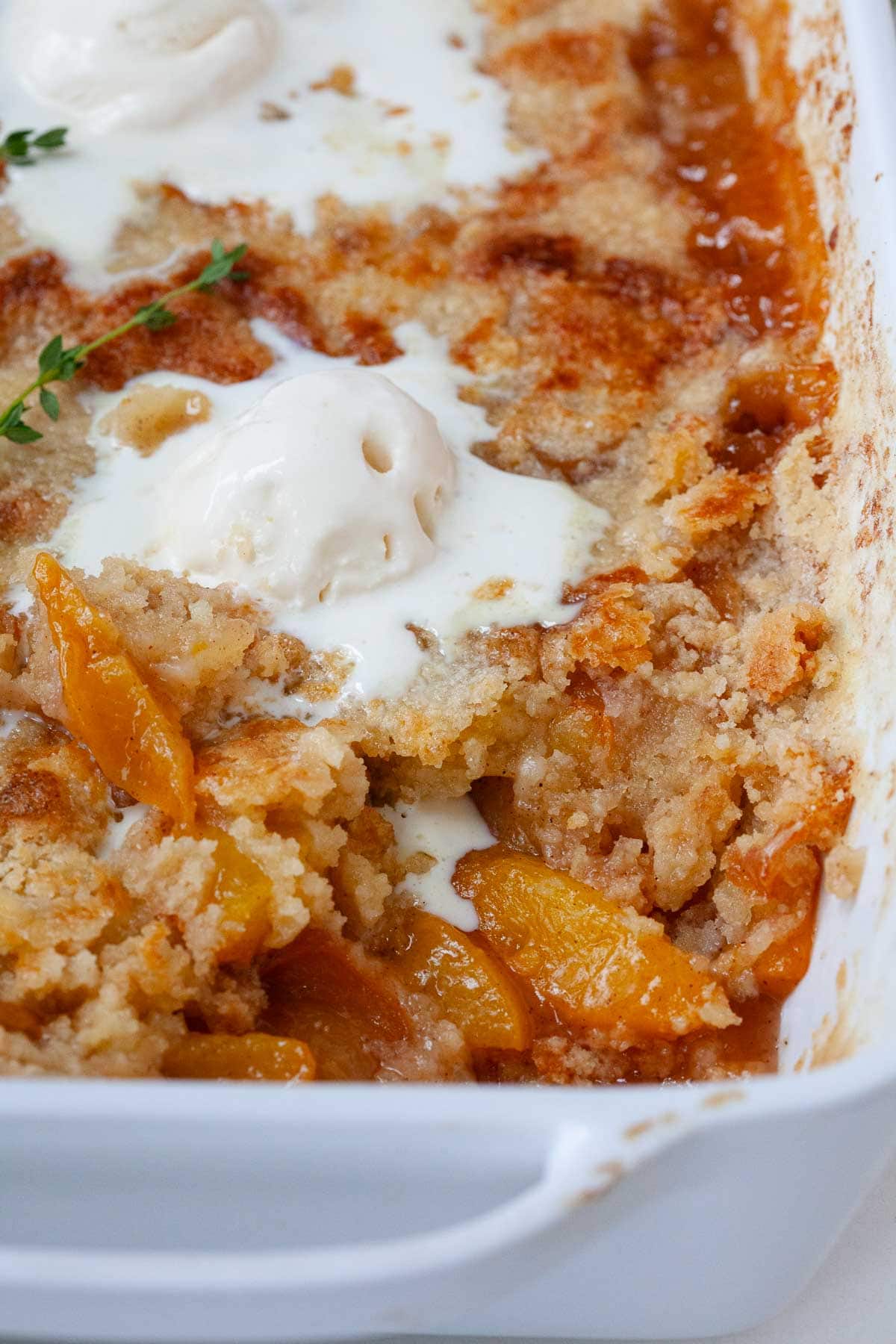 close up of cake mix peach cobbler in white baking dish with melting scoop of vanilla ice cream