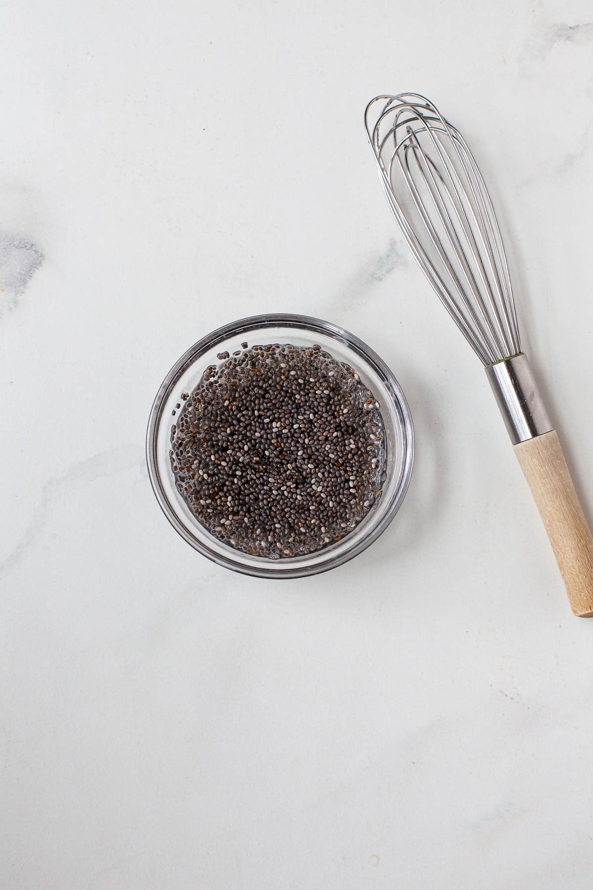 Chia seeds and water in small glass bowl on marble surface. Whisk on the right of the bowl. 