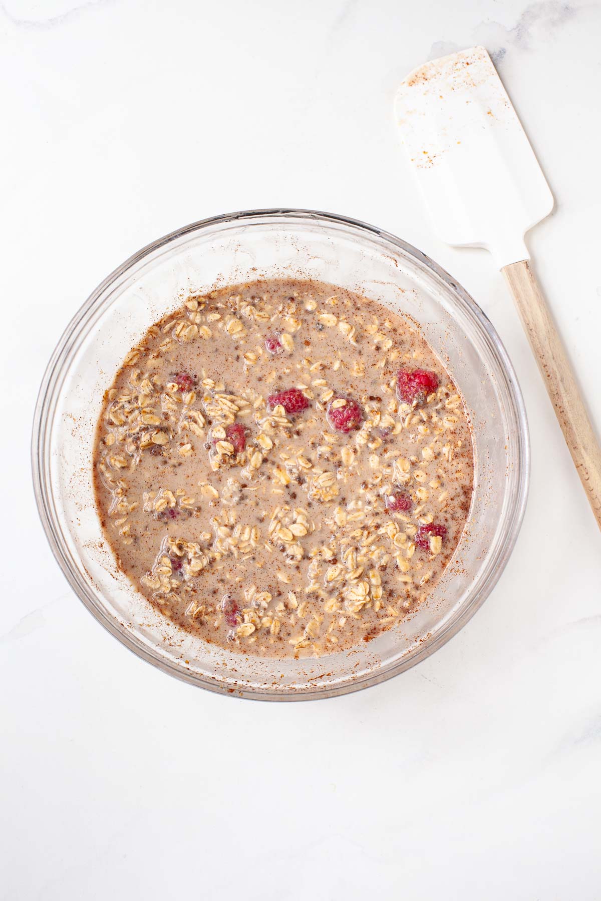 Raspberries folded into baked oatmeal mixture in glass bowl next to white spatula 