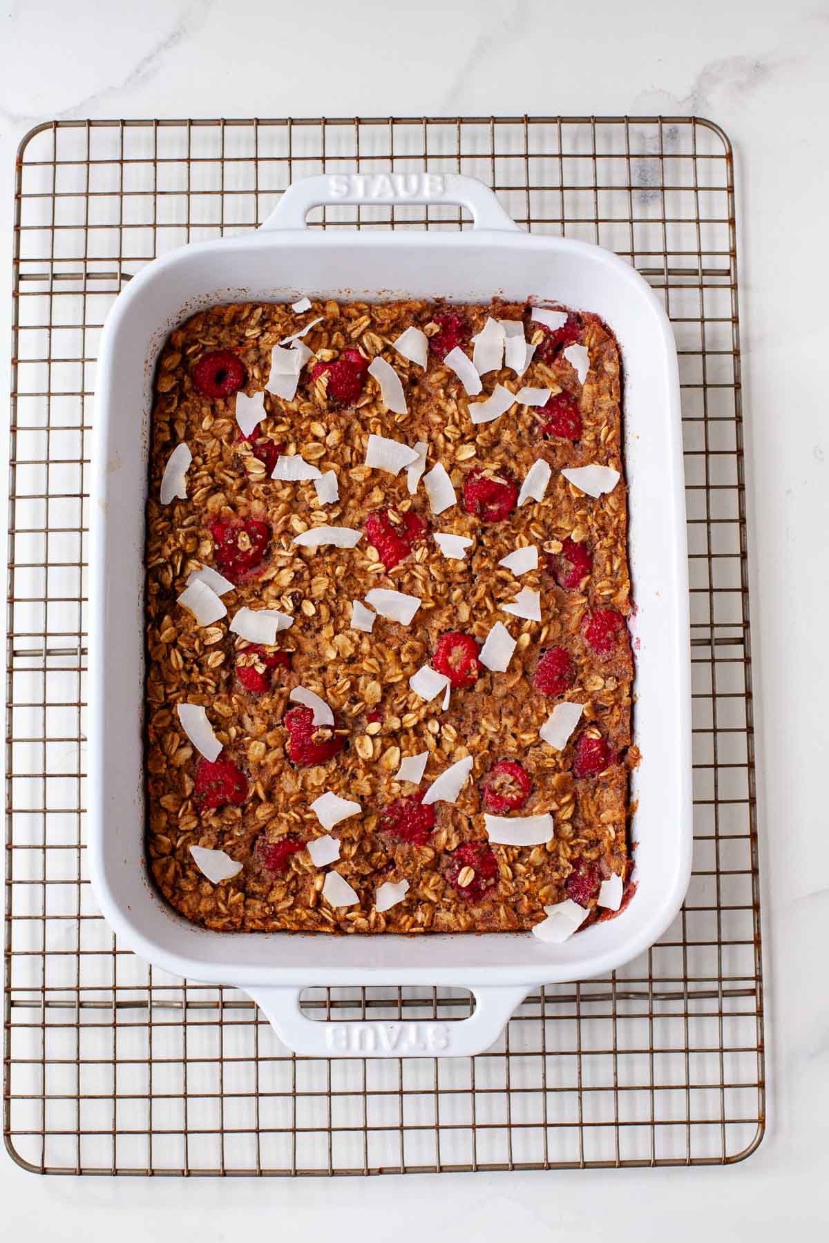 Baked vegan oatmeal topped with raspberries and coconut flakes in white ceramic baking dish on cooling rack. 